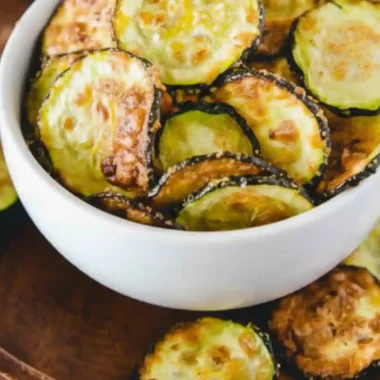 Crispy Air Fryer Zucchini Chips served in a white bowl on a wooden plate, showing golden-brown, thinly sliced zucchini rounds with a light, crunchy texture.