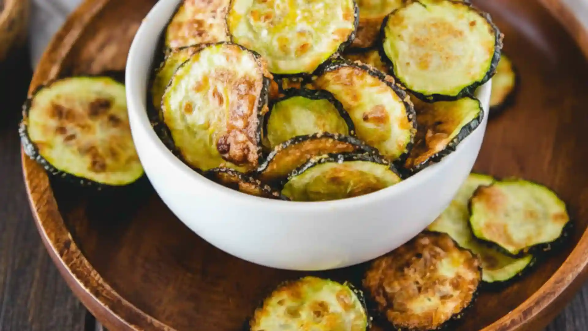 Crispy Air Fryer Zucchini Chips served in a white bowl on a wooden plate, showing golden-brown, thinly sliced zucchini rounds with a light, crunchy texture.