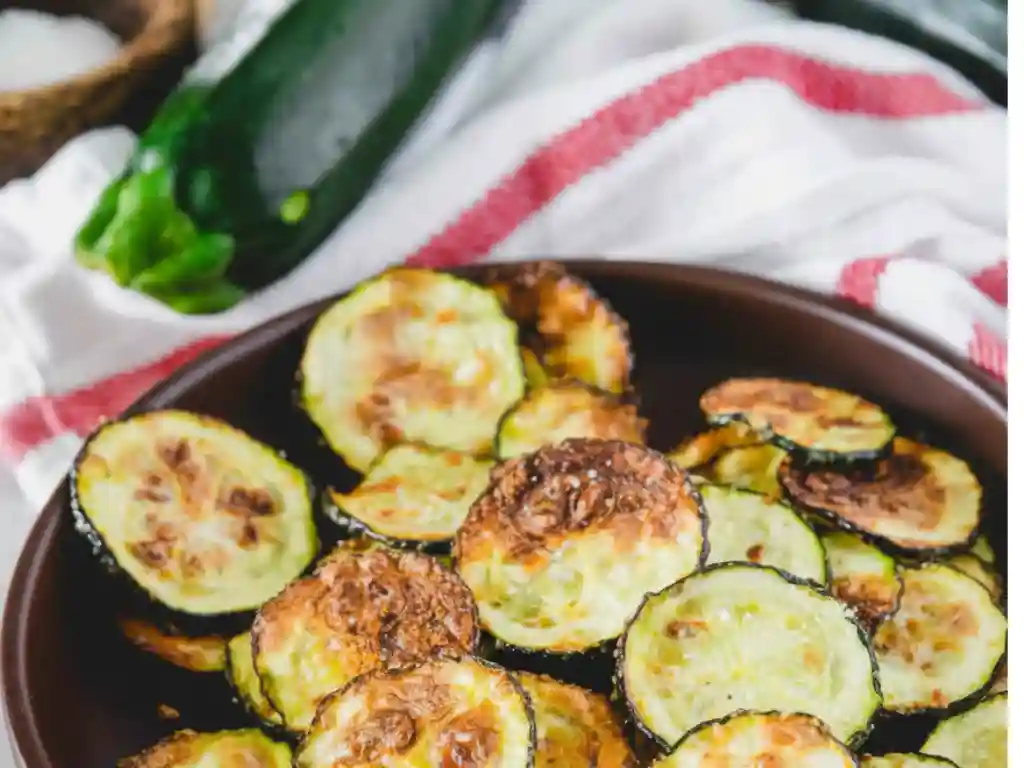 Crispy Air Fryer Zucchini Chips piled on a dark plate with golden-brown, thin zucchini rounds, a fresh zucchini and striped towel in the background
