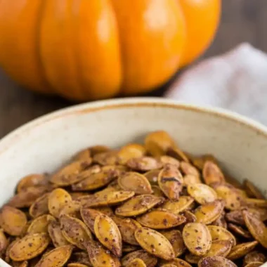 A bowl of savory roasted pumpkin seeds with a whole pumpkin in the background, illustrating a savory roasted pumpkin seeds recipe.