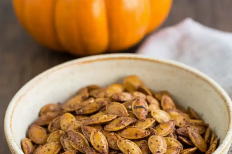A bowl of savory roasted pumpkin seeds with a whole pumpkin in the background, illustrating a savory roasted pumpkin seeds recipe.