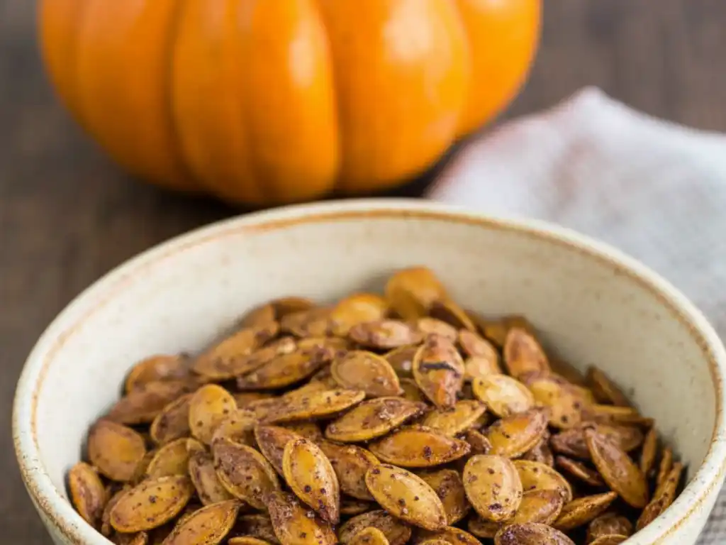 A bowl of savory roasted pumpkin seeds with a whole pumpkin in the background, illustrating a savory roasted pumpkin seeds recipe.