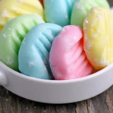 Amazing Cream Cheese Mints: Simple & Delicious 3 A small white bowl filled with homemade pastel cream cheese mints in pink, blue, green, and yellow, sitting on a rustic wooden surface.