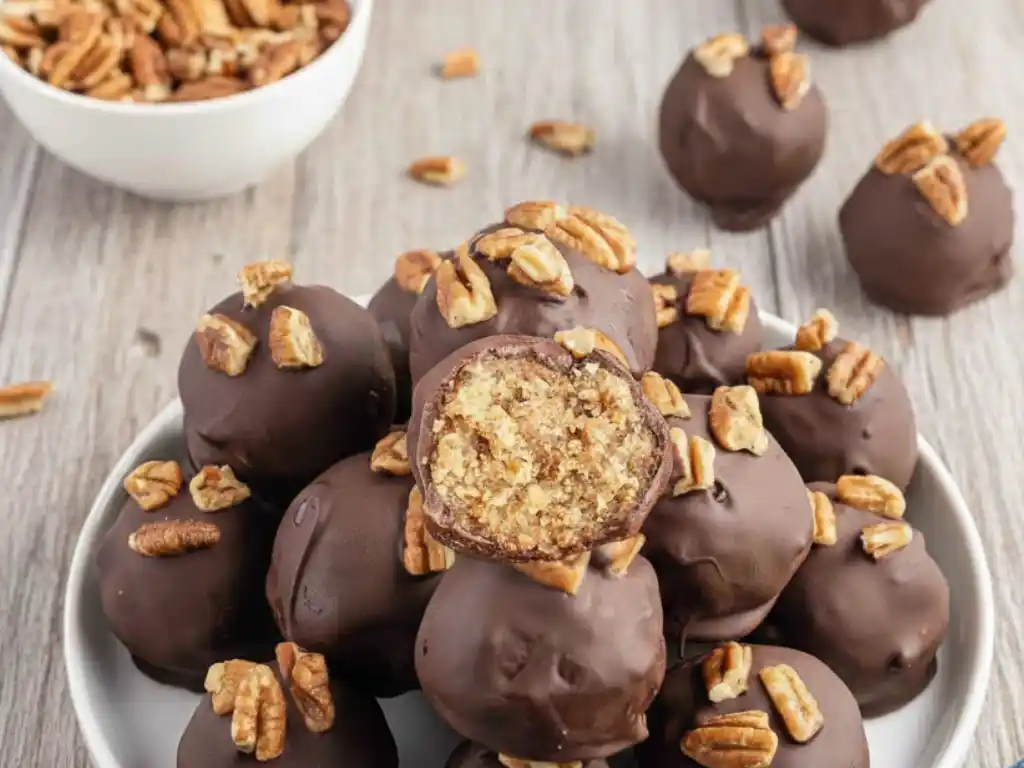 A plate of chocolate-covered pecan pie balls, with one cut in half to show the rich, crumbly pecan pie filling inside, and topped with chopped pecans.