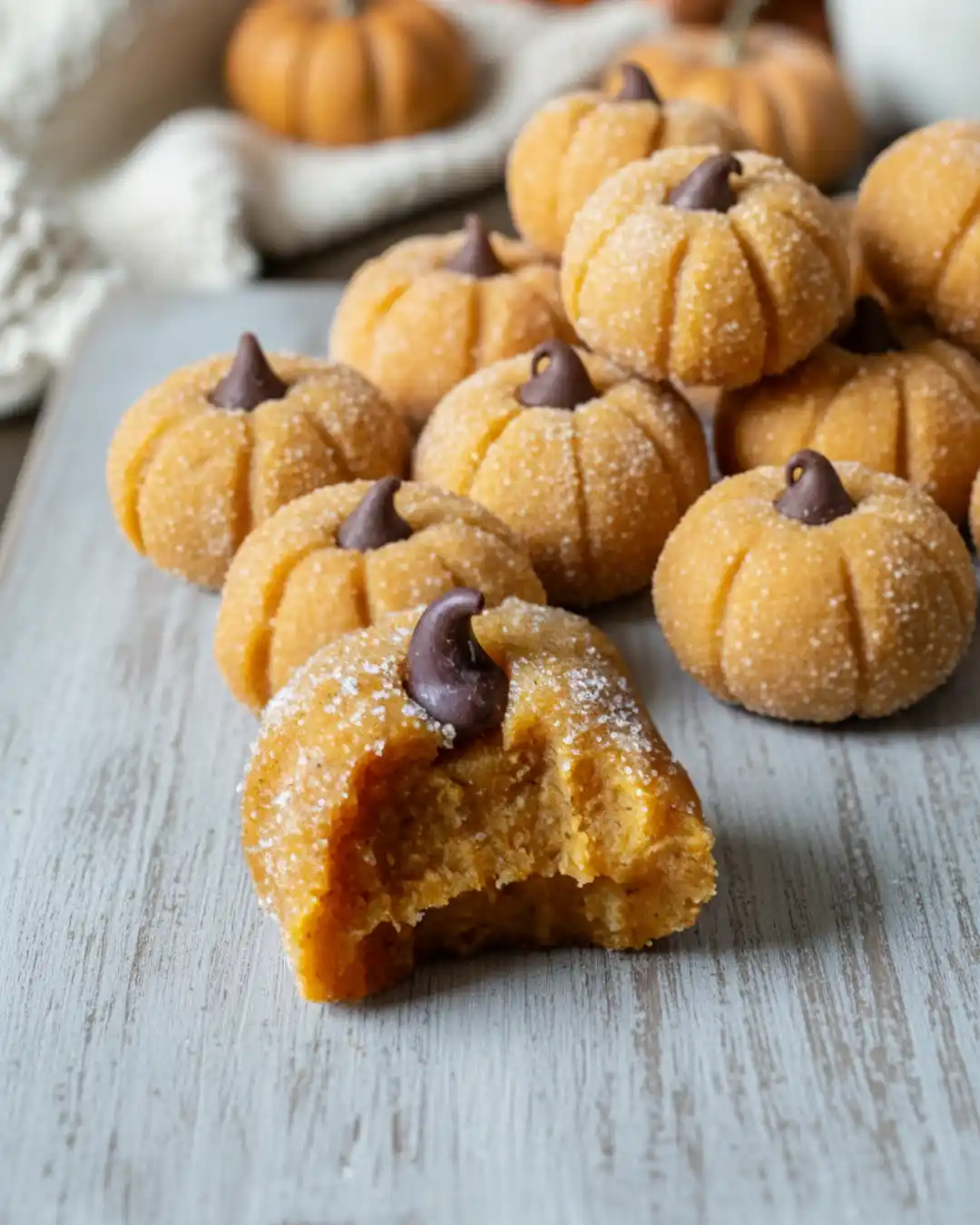 A collection of pumpkin cheesecake truffles on a wooden board, with one in the front that has a bite taken out to reveal the creamy orange pumpkin cheesecake filling inside.