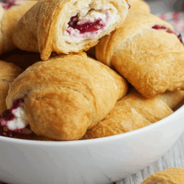Amazing Cranberry Cream Cheese Crescent Bites 3 A white bowl filled with golden-brown cranberry cream cheese crescent bites, with one broken open on top to reveal the warm cream cheese and cranberry filling.