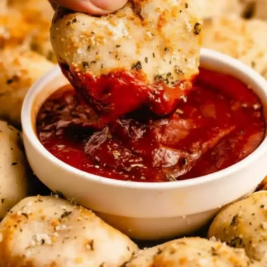 An array of golden-brown pizza bites, seasoned with herbs and parmesan, surrounding a small bowl of marinara dipping sauce topped with Italian seasoning.