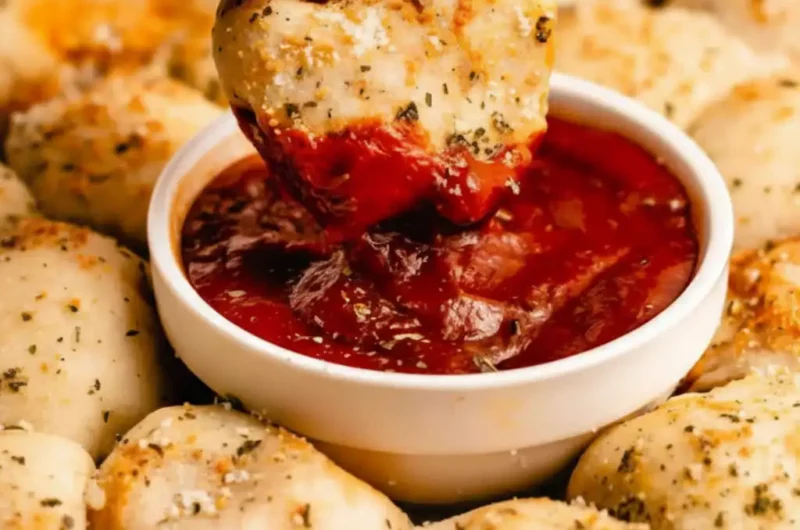 An array of golden-brown pizza bites, seasoned with herbs and parmesan, surrounding a small bowl of marinara dipping sauce topped with Italian seasoning.