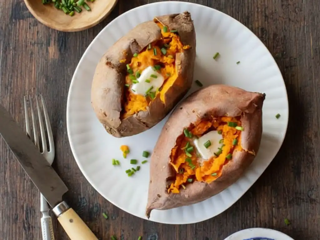 Amazing Baked Sweet Potato Oven Recipe 2 Two perfectly baked sweet potatoes split open on a white plate, topped with melting butter and fresh chives, ready to be served on a wooden table.