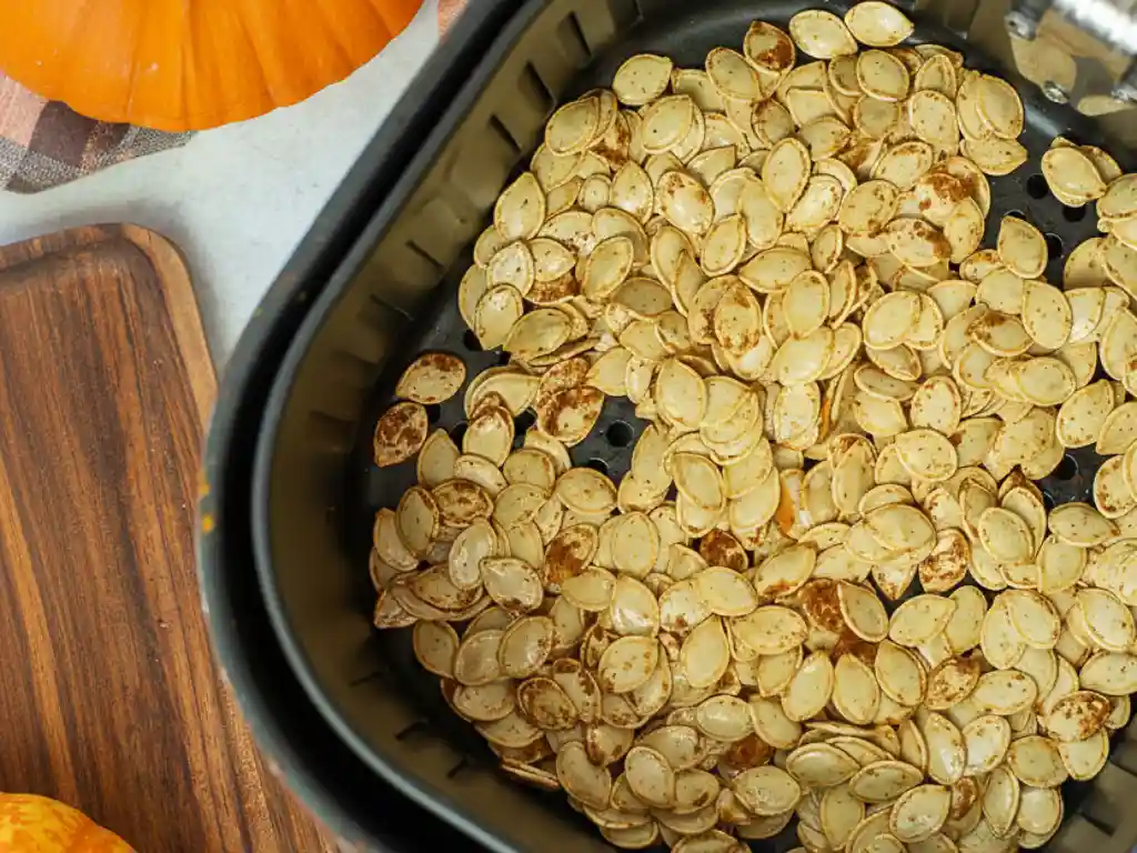 An overhead view of seasoned raw pumpkin seeds in an air fryer basket, ready to be cooked using an air fryer pumpkin seeds recipe, with a whole pumpkin and wooden board in the background
