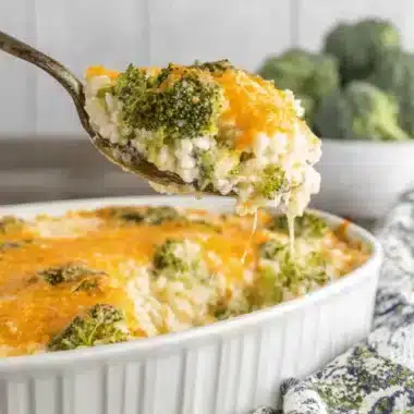 Wonderful Broccoli Cheese Rice Casserole 3 A spoonful of creamy, cheesy Broccoli Cheese Rice Casserole with stringy melted cheddar being lifted from a white baking dish, showing vibrant green broccoli and white rice.