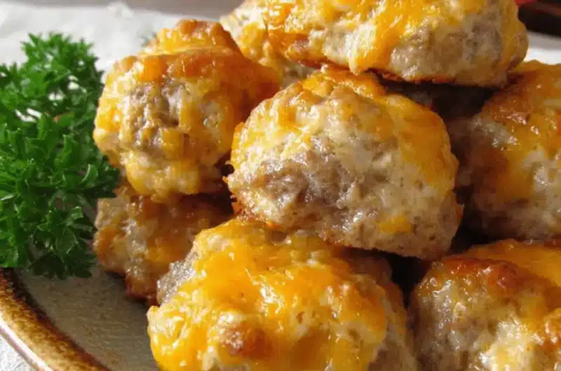 Close-up of golden-brown Cream Cheese Sausage Balls piled on a plate, generously topped with melted orange cheddar cheese and garnished with fresh parsley.