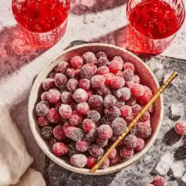 Vibrant flat lay of glistening sugared cranberries in a rustic ceramic bowl, flanked by refreshing red drinks and scattered ice.