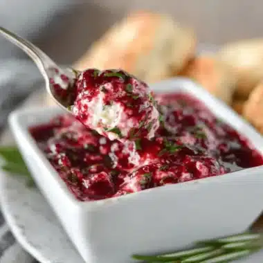 Close-up of a spoonful of vibrant Cranberry Jalapeno Dip being lifted from a white serving dish, showcasing its creamy texture, red cranberry swirls, and green herb flecks.