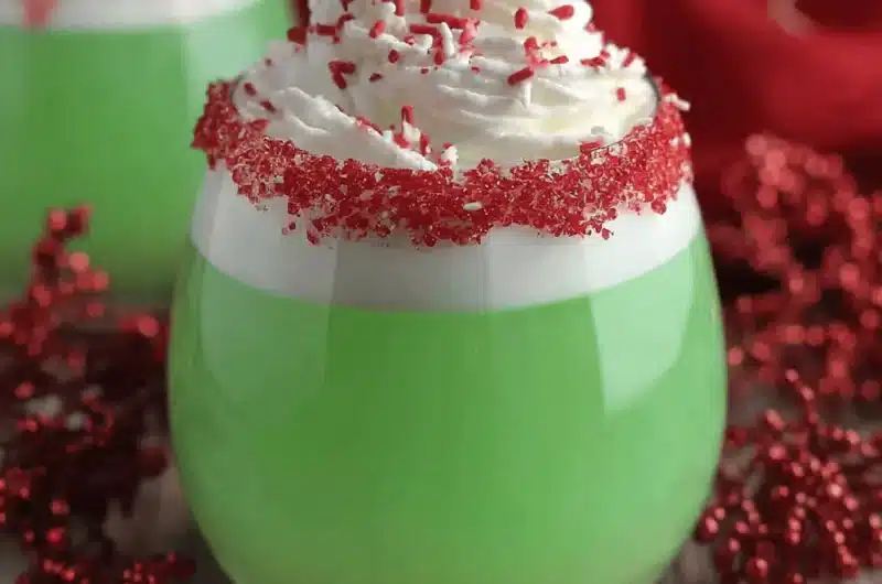 Vibrant Grinch Punch in a stemless glass, topped with whipped cream, red and white sprinkles, and a sparkling red sugar rim, set against a festive red background with holiday lights.