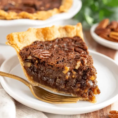 Ultimate Texas Chocolate Pecan Pie Recipe 3 A mouth-watering slice of Texas Chocolate Pecan Pie on a white plate with a gold fork, alongside a whole pie and scattered pecans in the background.