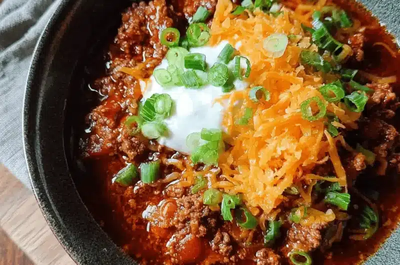 Hearty bowl of stovetop chili topped with sour cream, shredded cheese, and green onions on a rustic wooden surface.