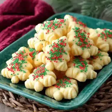 The Ultimate Reliable Spritz Cookies Recipe 3 Christmas tree-shaped spritz cookies with festive red and green sprinkles on a dark green plate.