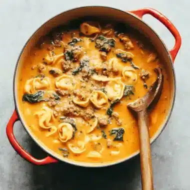 The Ultimate Creamy Tortellini Soup Recipe 4 Close-up of a rustic red Dutch oven filled with hearty tortellini soup, featuring cheese tortellini, Italian sausage, and greens.