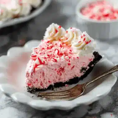 A delicious slice of Peppermint Pie with a dark chocolate cookie crust, pink peppermint filling, whipped cream rosettes, and crushed candy cane topping on a white scalloped plate with a fork.
