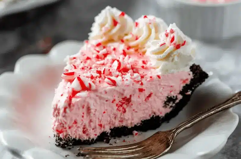A delicious slice of Peppermint Pie with a dark chocolate cookie crust, pink peppermint filling, whipped cream rosettes, and crushed candy cane topping on a white scalloped plate with a fork.