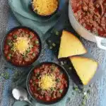 Overhead view of a hearty cowboy chili recipe with cornbread, topped with cheese and green onions on a rustic wooden surface.