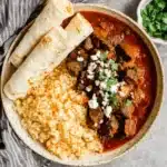 Rustic bowl of hearty chili colorado recipe served with fluffy rice and flatbread, garnished with cheese and fresh herbs.