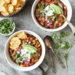 Two ceramic bowls of hearty Vegetarian Chili topped with sour cream, avocado, red onion, and cilantro, served with tortilla chips.