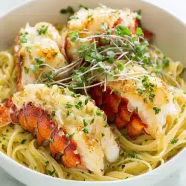 The Ultimate Butter Poached Lobster Recipe 3 Close-up of three succulent, split Butter Poached Lobster tails served over a bed of linguine pasta with fresh herbs and microgreens.