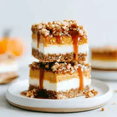 Stacked pumpkin cheesecake bars with caramel drizzle and streusel topping on a ceramic plate.