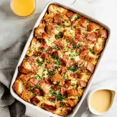 Savory baked Eggs Benedict Casserole with golden-brown bread, ham, and fresh chives, served with a side of hollandaise.