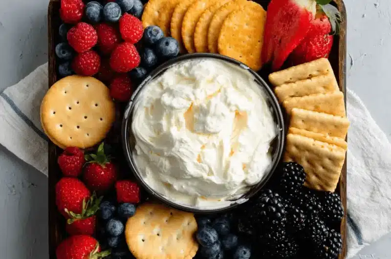 Flat-lay of a vibrant snack board centered around a bowl of creamy fruit dip with cream cheese, surrounded by fresh berries and assorted crackers.