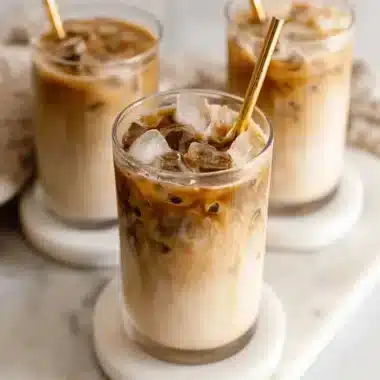 Three elegant glasses of layered iced coffee with swirling milk and dark espresso, featuring metallic gold straws on a bright white marble slab.