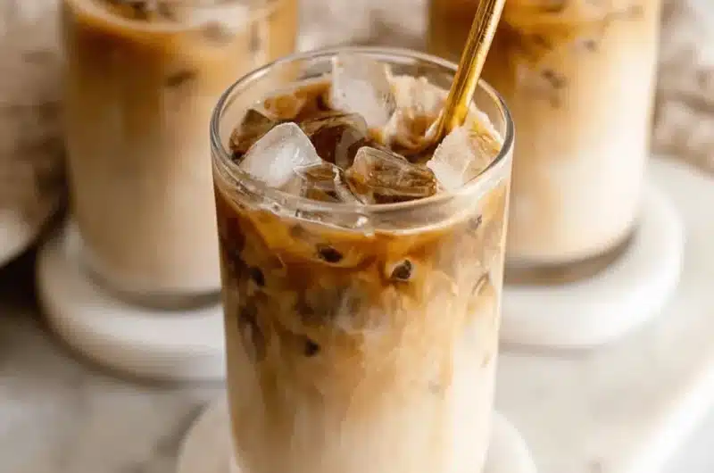 Three elegant glasses of layered iced coffee with swirling milk and dark espresso, featuring metallic gold straws on a bright white marble slab.