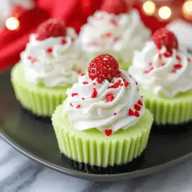 Surprising Grinch Mini Cheesecakes Recipe 3 Close-up of four festive Grinch Mini Cheesecakes featuring bright green filling, red heart sprinkles, and a dark crust on a matte black serving dish.
