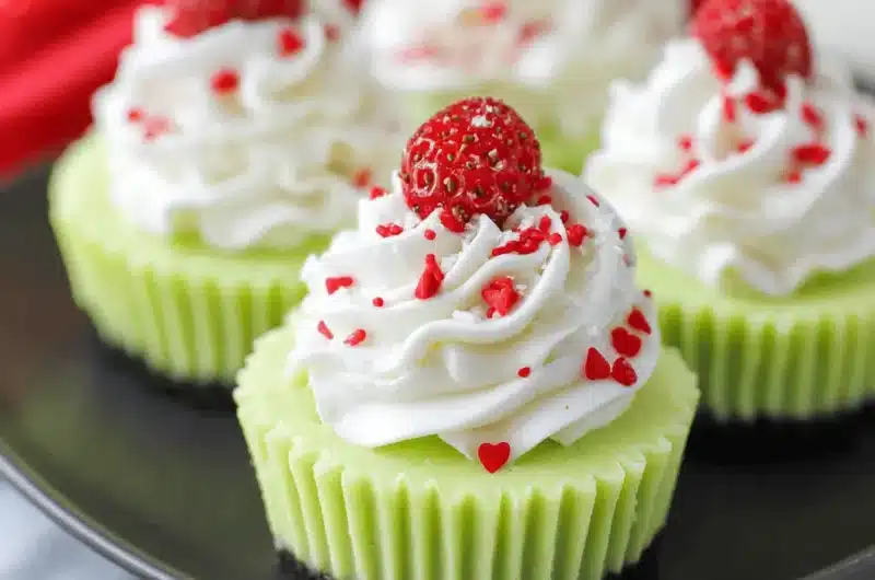 Close-up of four festive Grinch Mini Cheesecakes featuring bright green filling, red heart sprinkles, and a dark crust on a matte black serving dish.