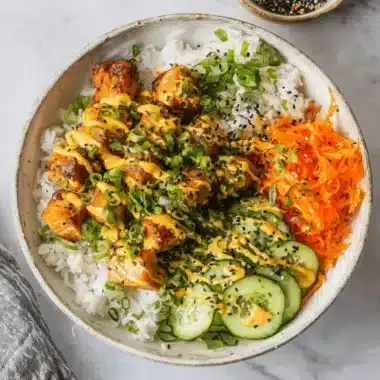 Overhead view of a delicious glazed salmon bowl with rice, shredded carrots, cucumber, spicy mayo, and black sesame seeds.