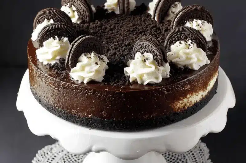 Detailed photo of a tall, decadent triple-layer oreo cheesecake topped with chocolate ganache, whipped cream swirls, and whole Oreo cookies on a white pedestal stand.