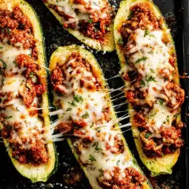 Four baked stuffed zucchini boats with stringy melted mozzarella cheese and savory meat filling on a dark tray.