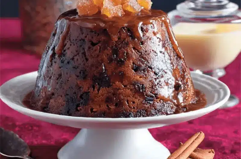 High-quality photo of a rich, sauce-covered Christmas Pudding topped with candied ginger, set on a pedestal stand against deep crimson velvet with cinnamon sticks and warm candlelight.
