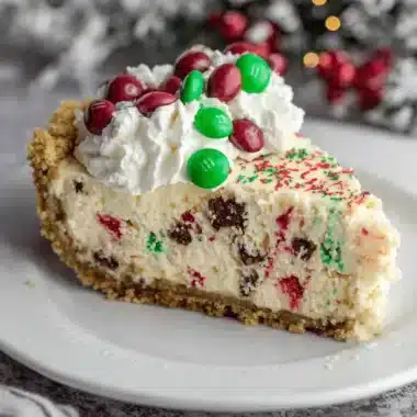 Slice of decadent christmas cheesecake topped with whipped cream, red and green M&Ms, and festive sprinkles.