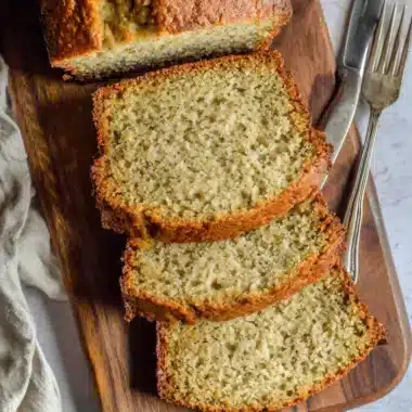 Overhead view of moist slices of freshly baked cake mix banana bread resting on a dark wooden cutting board with a vintage knife and fork.