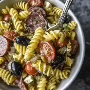 Hearty Italian-style rotini pasta salad with salami, cherry tomatoes, and fresh mozzarella being scooped with a silver spoon.