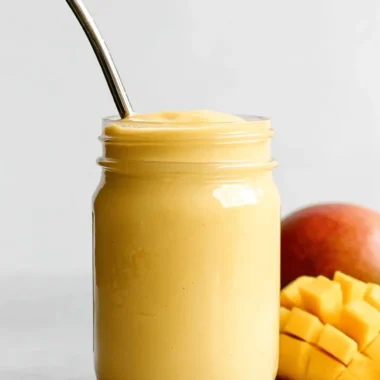 Close-up of a vibrant yellow mango smoothie in a mason jar, served with a reusable straw next to fresh sliced mango.