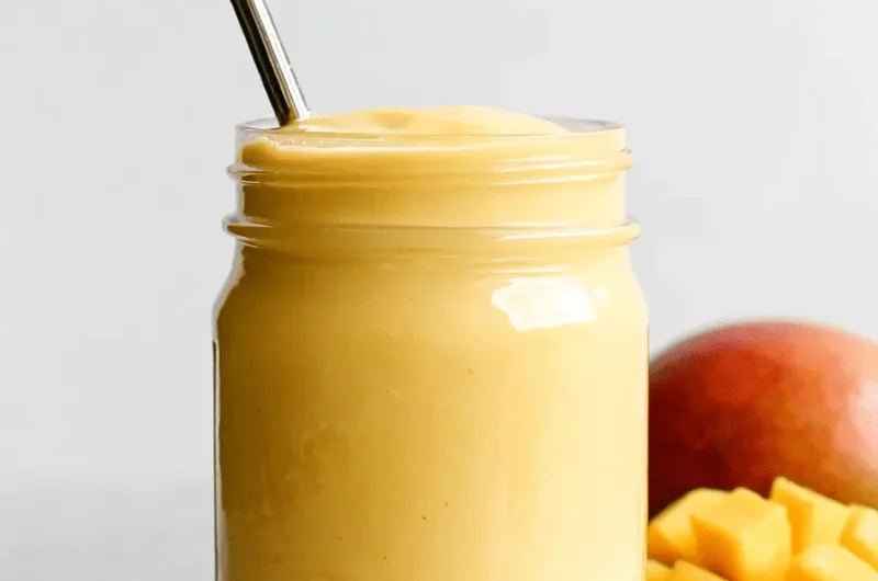 Close-up of a vibrant yellow mango smoothie in a mason jar, served with a reusable straw next to fresh sliced mango.