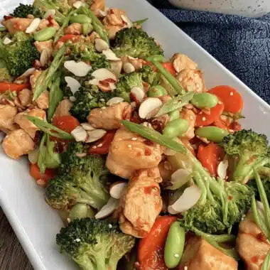 Appetizing image of glossy chicken stir fry with vegetables (broccoli, carrots, edamame) and sliced almonds on a white platter.