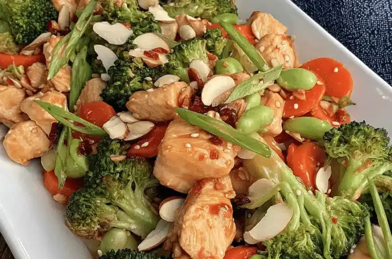 Appetizing image of glossy chicken stir fry with vegetables (broccoli, carrots, edamame) and sliced almonds on a white platter.