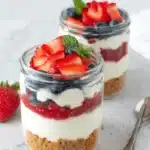 Two clear glass cheesecake jars showing layers of crust, cream, and red fruit compote, garnished with fresh berries and mint.