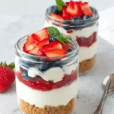 Simple & Amazing Cheesecake Jars Recipe 3 Two clear glass cheesecake jars showing layers of crust, cream, and red fruit compote, garnished with fresh berries and mint.