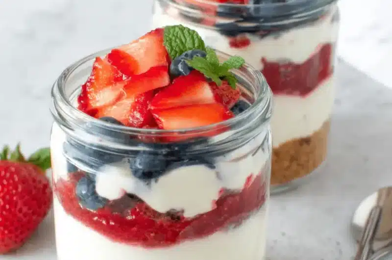 Two clear glass cheesecake jars showing layers of crust, cream, and red fruit compote, garnished with fresh berries and mint.